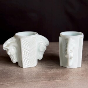 May include: Two white ceramic vases. One vase features two horse heads on either side, with a chevron pattern on the front. The other vase has a single horse head on the front. Both vases have a light blue interior.