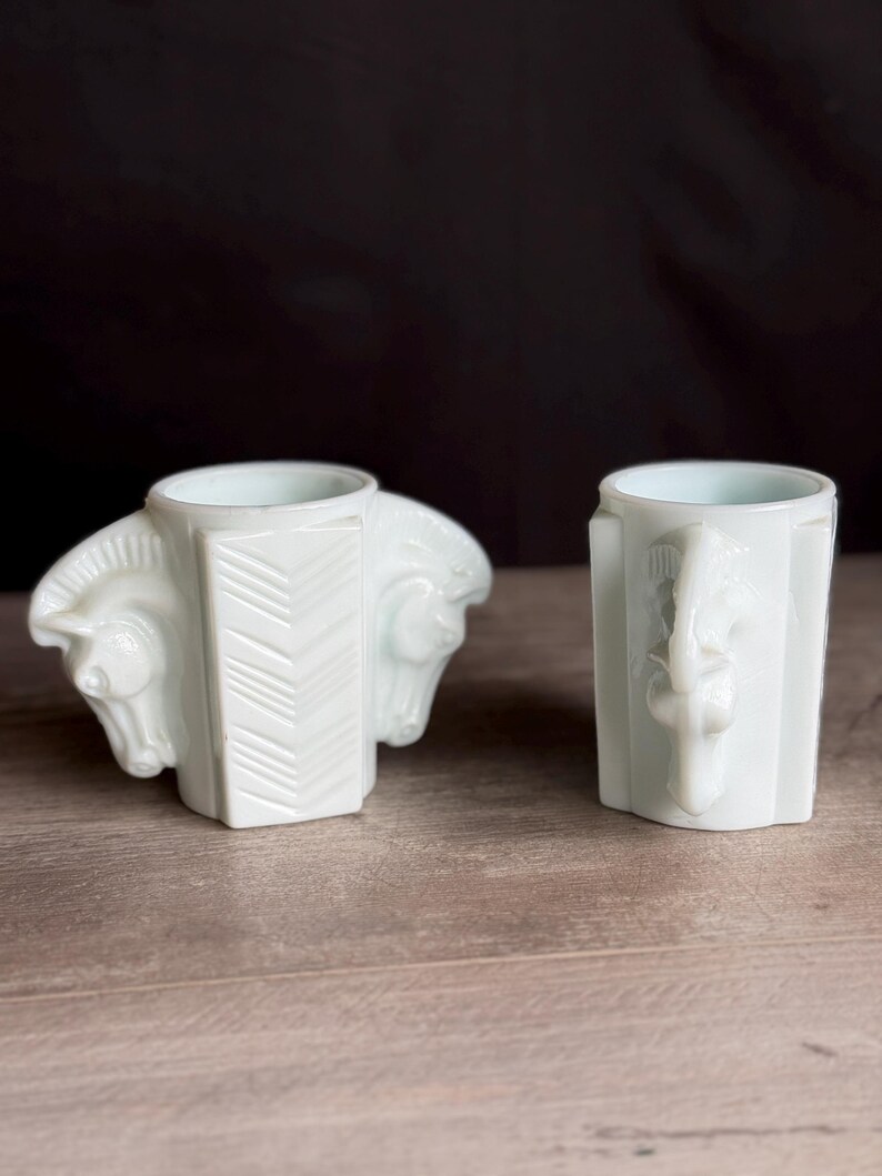 May include: Two white ceramic vases. One vase features two horse heads on either side, with a chevron pattern on the front. The other vase has a single horse head on the front. Both vases have a light blue interior.