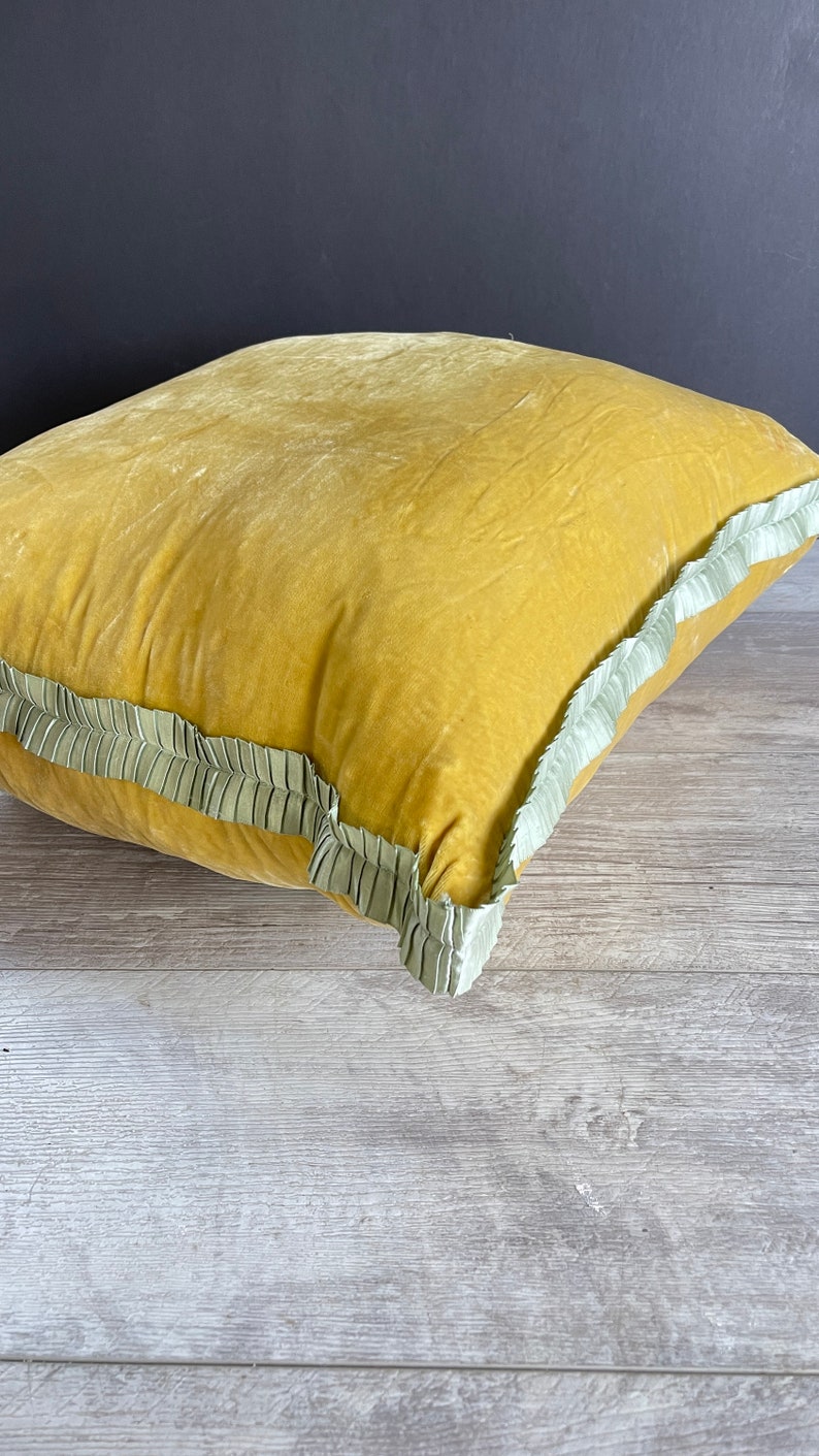 K&ouml;nnte beinhalten: Ein senfgelbes Samtkissen mit einem hellgr&uuml;nen R&uuml;schenbesatz.