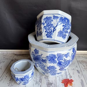 May include: Three white and blue ceramic planters with a grape vine design. The planters are octagonal and vary in size, with the largest one at the bottom and the smallest one on the left. A red leaf is on the wooden surface.