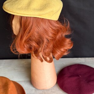 Puede incluir: Una gorra de peri&oacute;dico amarilla se muestra sobre una cabeza de maniqu&iacute; con cabello rojo. Tambi&eacute;n se exhiben una boina granate, un fedora naranja y dos hojas coloridas. Los art&iacute;culos est&aacute;n sobre una superficie de madera clara con un fondo negro.