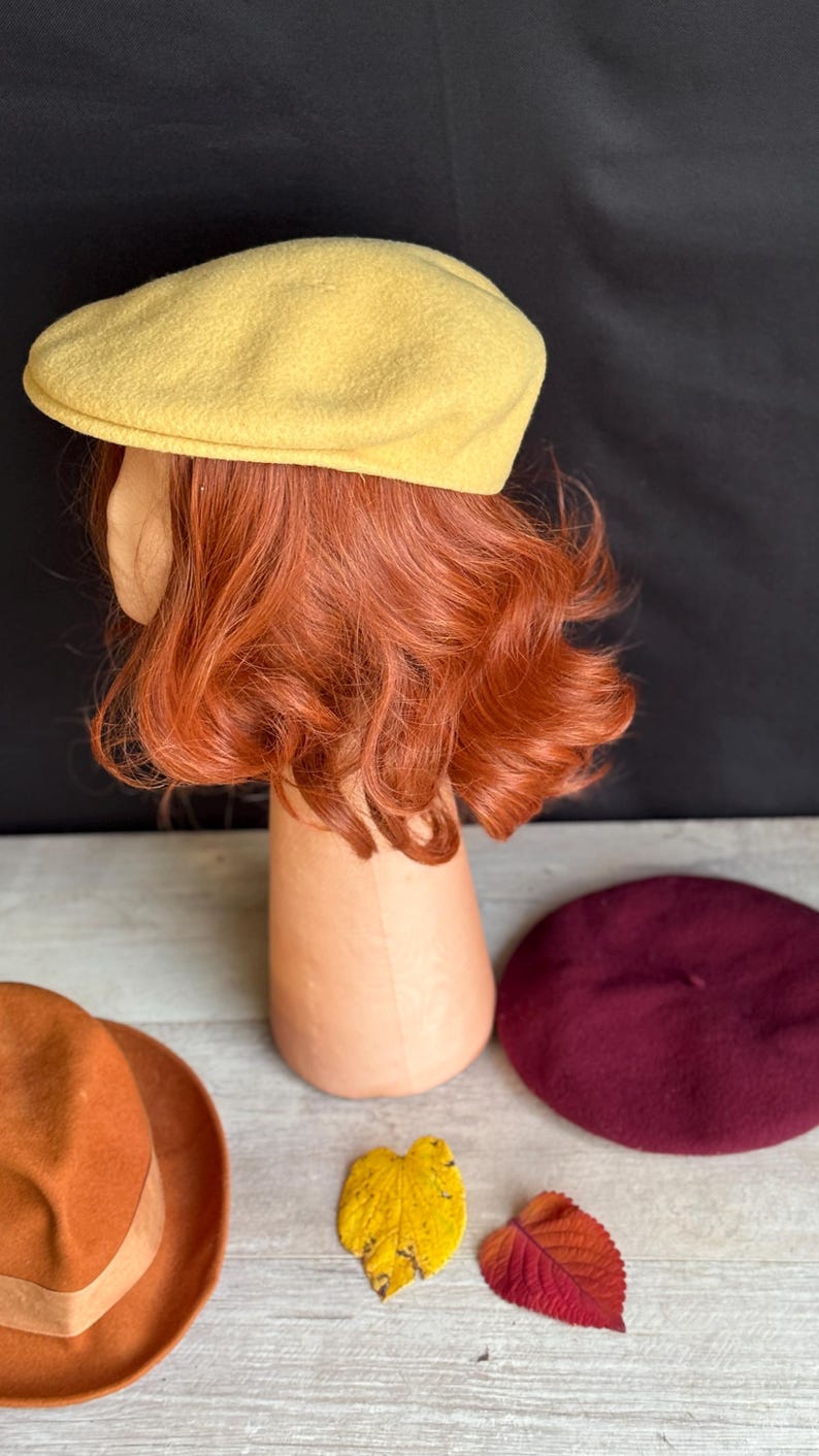 Puede incluir: Una gorra de peri&oacute;dico amarilla se muestra sobre una cabeza de maniqu&iacute; con cabello rojo. Tambi&eacute;n se exhiben una boina granate, un fedora naranja y dos hojas coloridas. Los art&iacute;culos est&aacute;n sobre una superficie de madera clara con un fondo negro.