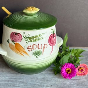 May include: A vintage soup tureen with a green lid and a matching spoon. The ceramic bowl is decorated with painted vegetables and the word "soup". Pink and orange flowers and greenery are next to the tureen.