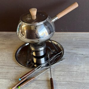 May include: A vintage fondue set with a stainless steel pot, wooden handle, and lid. The set includes a burner, a tray, and fondue forks with colored tips. The pot is approximately 6 inches in diameter.