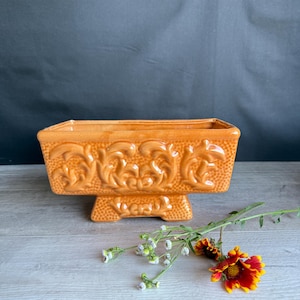 May include: A rectangular orange ceramic planter with a glossy finish, featuring raised scrollwork patterns on the front and a textured base. The planter is accompanied by a small sprig of wildflowers with red and yellow petals and green stems.