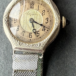 May include: A vintage silver-toned wristwatch with a mesh band. The watch face is gold-colored with black numerals and hands. The text "WESTPOINTER" and "SWISS" are visible on the watch face. The watch has an ornate, engraved bezel.