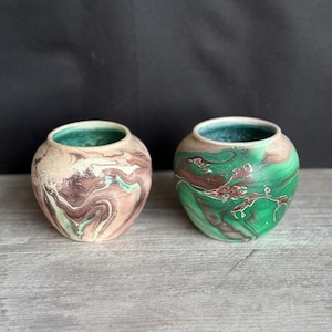 May include: Two small, round ceramic vases with a marbled design. One vase is primarily beige and brown with teal accents, while the other is green and brown with white accents. Both have a teal interior and sit on a wooden surface.