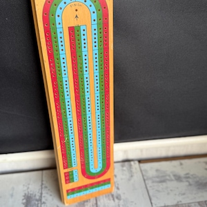 May include: Wooden cribbage board with a colorful track. The board features a U-shaped track with red, green, and blue lanes, and is marked with numbers and the text "THE U.S. PLAYING CARD CO." at the top. The board is rectangular and has a light wood finish.