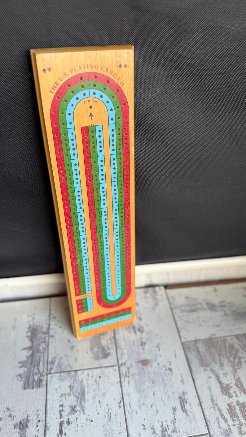 May include: Wooden cribbage board with a colorful track. The board features a U-shaped track with red, green, and blue lanes, and is marked with numbers and the text "THE U.S. PLAYING CARD CO." at the top. The board is rectangular and has a light wood finish.