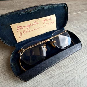May include: Gold-tone metal eyeglasses with round lenses in a dark blue velvet-lined hard case. A cream-colored paper insert inside the case features handwritten red ink text that reads: Margarite firt glasses - 1953.