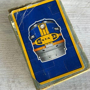 May include: Vintage playing card box featuring a blue background with a yellow and silver train illustration. The text "SANTA FE" is displayed on the train. The box has a gold border and shows signs of wear.