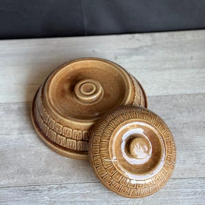May include: A brown ceramic serving dish with a lid. The dish and lid are a matching shade of brown and have a textured, brick-like pattern around the sides. The lid has a central knob for easy handling.
