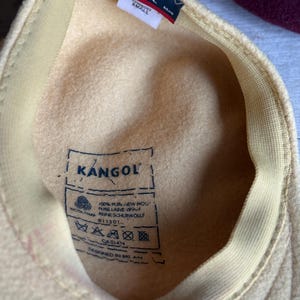 Puede incluir: Sombrero Kangol amarillo claro, visto desde el interior, con un interior color crema y un borde cosido. El sombrero presenta una peque&ntilde;a etiqueta con el logotipo de Kangol y la palabra "SMALL". El sombrero es de lana.