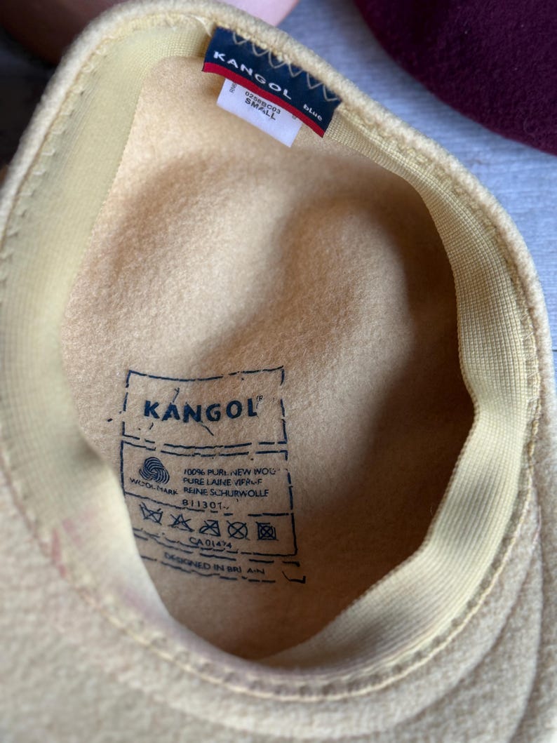 Puede incluir: Sombrero Kangol amarillo claro, visto desde el interior, con un interior color crema y un borde cosido. El sombrero presenta una peque&ntilde;a etiqueta con el logotipo de Kangol y la palabra "SMALL". El sombrero es de lana.