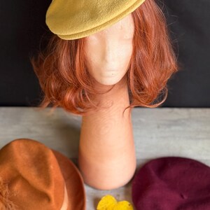 Puede incluir: Un boina amarillo mostaza se asienta sobre una cabeza de maniqu&iacute; con cabello casta&ntilde;o rojizo. Un sombrero fedora naranja y una boina burdeos se exhiben debajo. Hojas de oto&ntilde;o en amarillo y rojo a&ntilde;aden un toque estacional.
