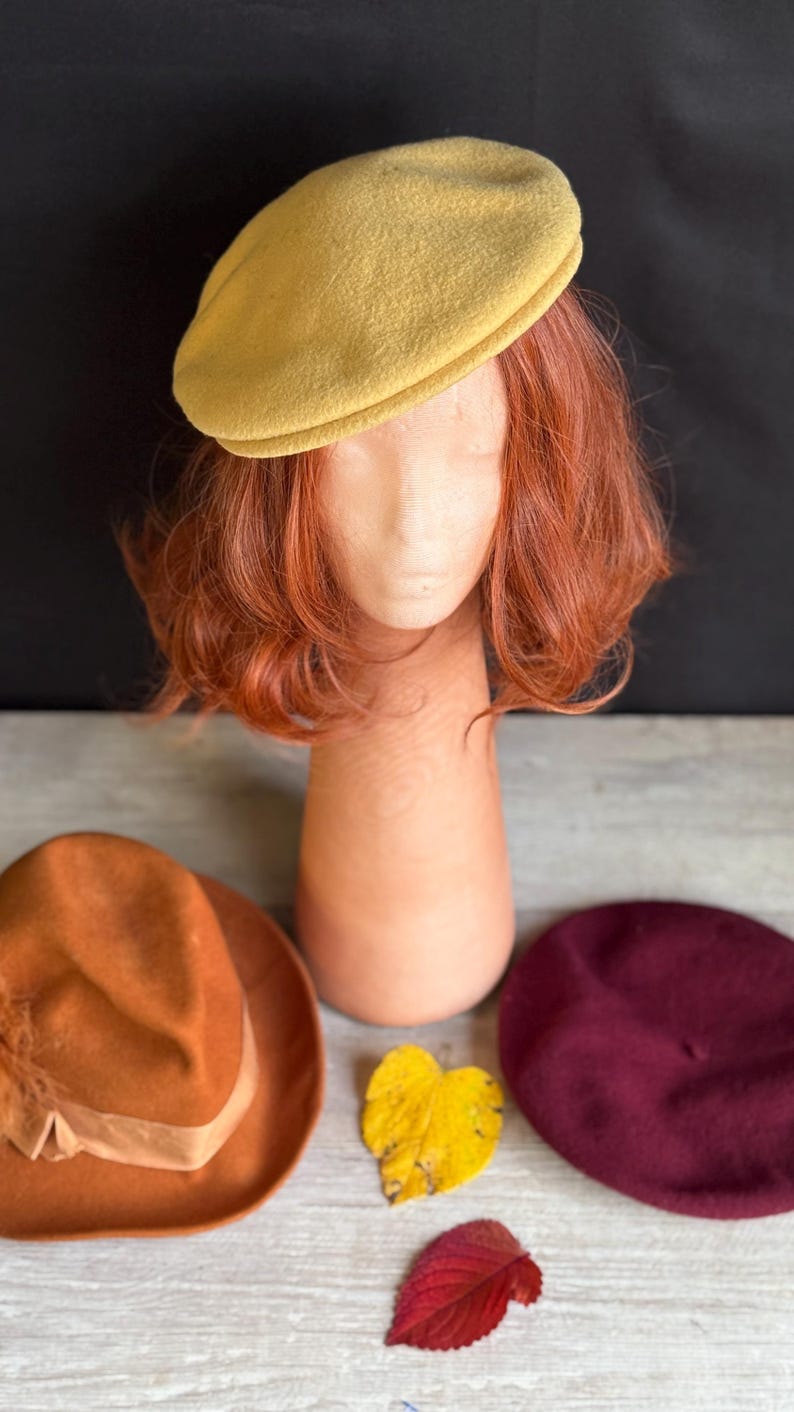 Puede incluir: Un boina amarillo mostaza se asienta sobre una cabeza de maniqu&iacute; con cabello casta&ntilde;o rojizo. Un sombrero fedora naranja y una boina burdeos se exhiben debajo. Hojas de oto&ntilde;o en amarillo y rojo a&ntilde;aden un toque estacional.