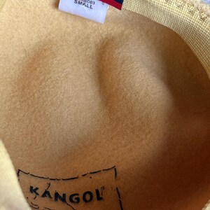 Puede incluir: Primer plano de un sombrero Kangol amarillo. El sombrero tiene una etiqueta azul con el nombre de la marca y la palabra "blue". El sombrero est&aacute; etiquetado como "SMALL". El interior del sombrero tiene una etiqueta Kangol.