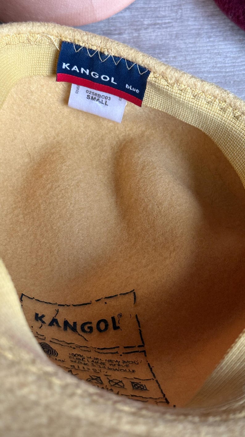 Puede incluir: Primer plano de un sombrero Kangol amarillo. El sombrero tiene una etiqueta azul con el nombre de la marca y la palabra "blue". El sombrero est&aacute; etiquetado como "SMALL". El interior del sombrero tiene una etiqueta Kangol.
