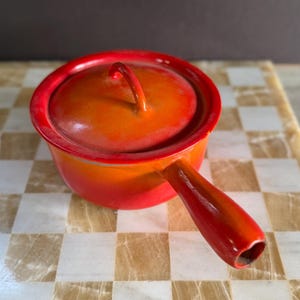 May include: A small, round, orange and red enameled cast iron saucepan with a lid and a single handle. The cookware sits on a checkered marble surface. The handle and rim are a darker red.