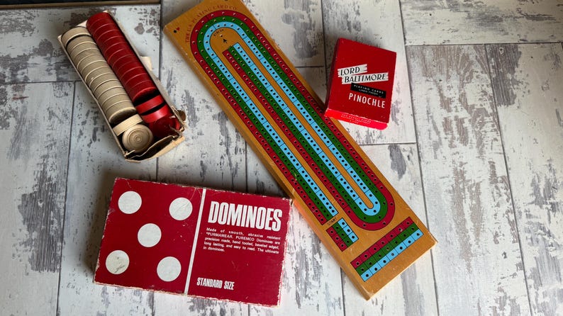May include: A collection of vintage board games, including a cribbage board, a deck of "Lord Baltimore Pinochle" playing cards, a box of dominoes, and a box of cribbage pegs.
