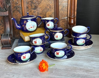 May include: A cobalt blue porcelain tea set featuring gold trim and hand-painted illustrations of figures in red clothing. The set includes a teapot, a creamer, a lidded sugar bowl, and four teacups with matching saucers, all decorated with gold accents.