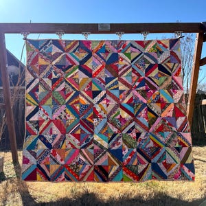 May include: A colorful patchwork quilt with a geometric design, featuring a variety of fabrics in shades of red, blue, green, and yellow. The quilt is displayed outdoors, hanging from a wooden frame, with a clear blue sky in the background.