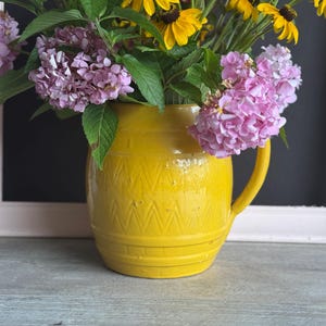 May include: A yellow ceramic pitcher filled with a vibrant bouquet of flowers. The flowers include yellow coneflowers, pink hydrangeas, and other colorful blooms. The pitcher has a textured design and a handle, sitting on a wooden surface.