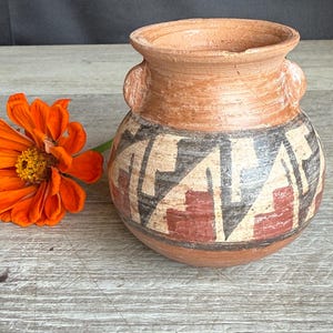 May include: A small, round, terracotta-colored pottery vase with a geometric design in black, tan, and red. The vase has two small handles and an orange flower is next to it.
