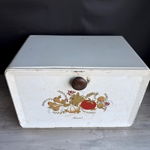 May include: A vintage white bread box with a wooden knob and a decorative illustration of vegetables. The illustration includes a red tomato, garlic, and mushrooms. The word "Bread" is printed below the illustration.