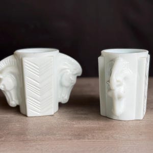May include: Two white art deco style vases. One vase features two horse heads on either side, with a geometric design in the center. The other vase has a seahorse design on the front. Both vases are on a wooden surface.