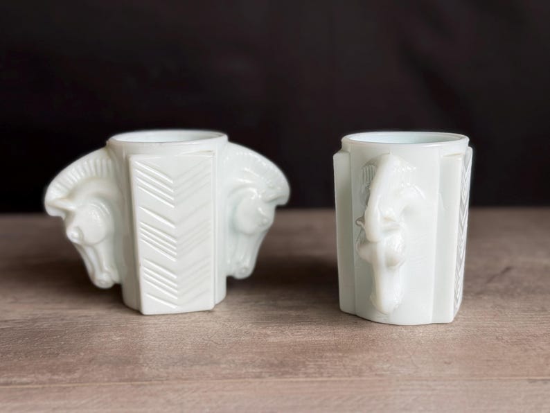 May include: Two white art deco style vases. One vase features two horse heads on either side, with a geometric design in the center. The other vase has a seahorse design on the front. Both vases are on a wooden surface.