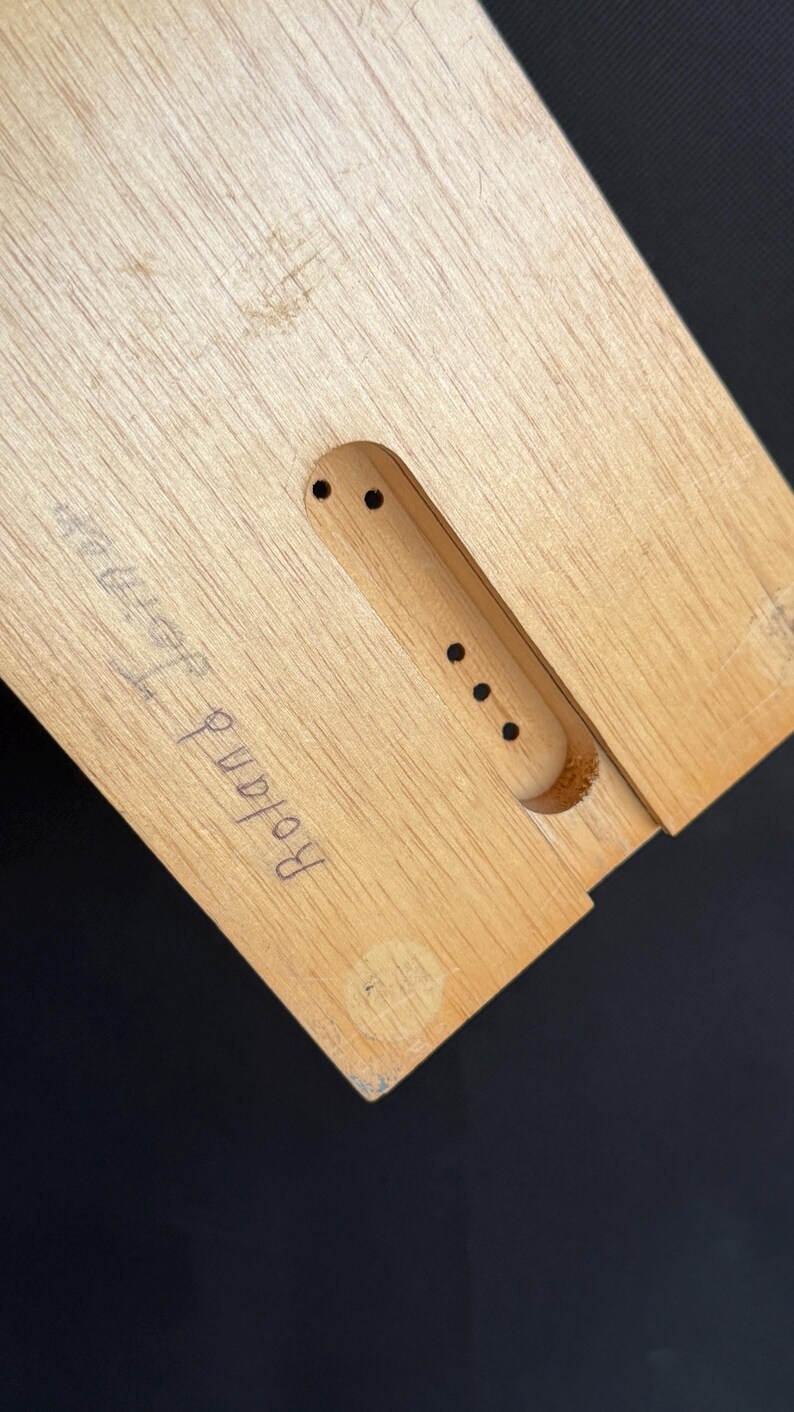 May include: Close-up of a light-colored wooden object with a rectangular cutout and several small holes. The name "Roland Herman" is handwritten in blue ink on the wood. The background is black.