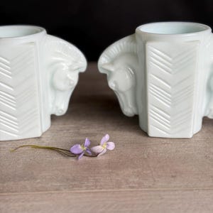 May include: Two white, Art Deco-style bookends, each featuring a horse head design. The bookends have a geometric pattern on the sides. A sprig of purple flowers rests on the wooden surface between the bookends.