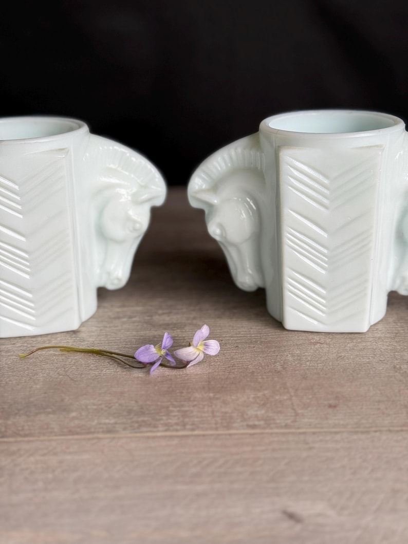 May include: Two white, Art Deco-style bookends, each featuring a horse head design. The bookends have a geometric pattern on the sides. A sprig of purple flowers rests on the wooden surface between the bookends.
