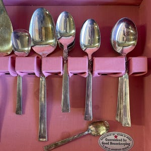 May include: A set of silver-plated Kingswood by Empire Crafts spoons in a pink box. The spoons are arranged in a row and are all the same size. The box has a label that says "Replacement or Refund of Money Guaranteed by Good Housekeeping if not as advertised therein."