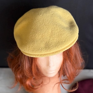 Puede incluir: Gorra de estilo newsboy de color amarillo mostaza. El sombrero est&aacute; hecho de una tela suave y texturizada y tiene una corona redondeada y una peque&ntilde;a visera r&iacute;gida. La gorra se exhibe en una cabeza de maniqu&iacute; con cabello rojo.