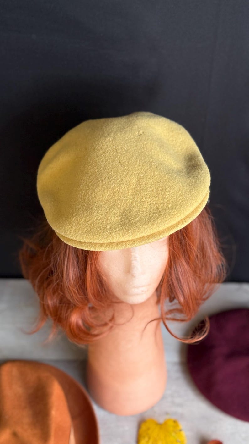 Puede incluir: Gorra de estilo newsboy de color amarillo mostaza. El sombrero est&aacute; hecho de una tela suave y texturizada y tiene una corona redondeada y una peque&ntilde;a visera r&iacute;gida. La gorra se exhibe en una cabeza de maniqu&iacute; con cabello rojo.