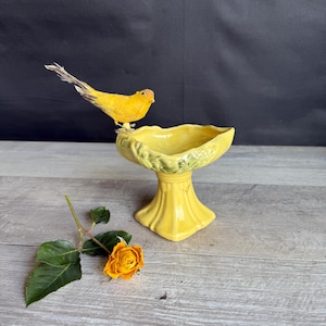 May include: A yellow ceramic pedestal bowl with a decorative rim, topped with a yellow bird figurine. A dried orange rose and green leaves are on the wooden surface. The bowl is a decorative item.
