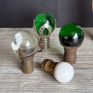 May include: A collection of decorative finials with various designs. Includes a clear glass ball finial, a green glass ball finial with silver accents, a green and black glass ball finial, and a white ball finial.