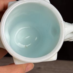 May include: A white ceramic mug with a light blue interior. The mug has a square base and a rounded rim. The handle is white and appears to be sculpted. The mug is set against a dark background.