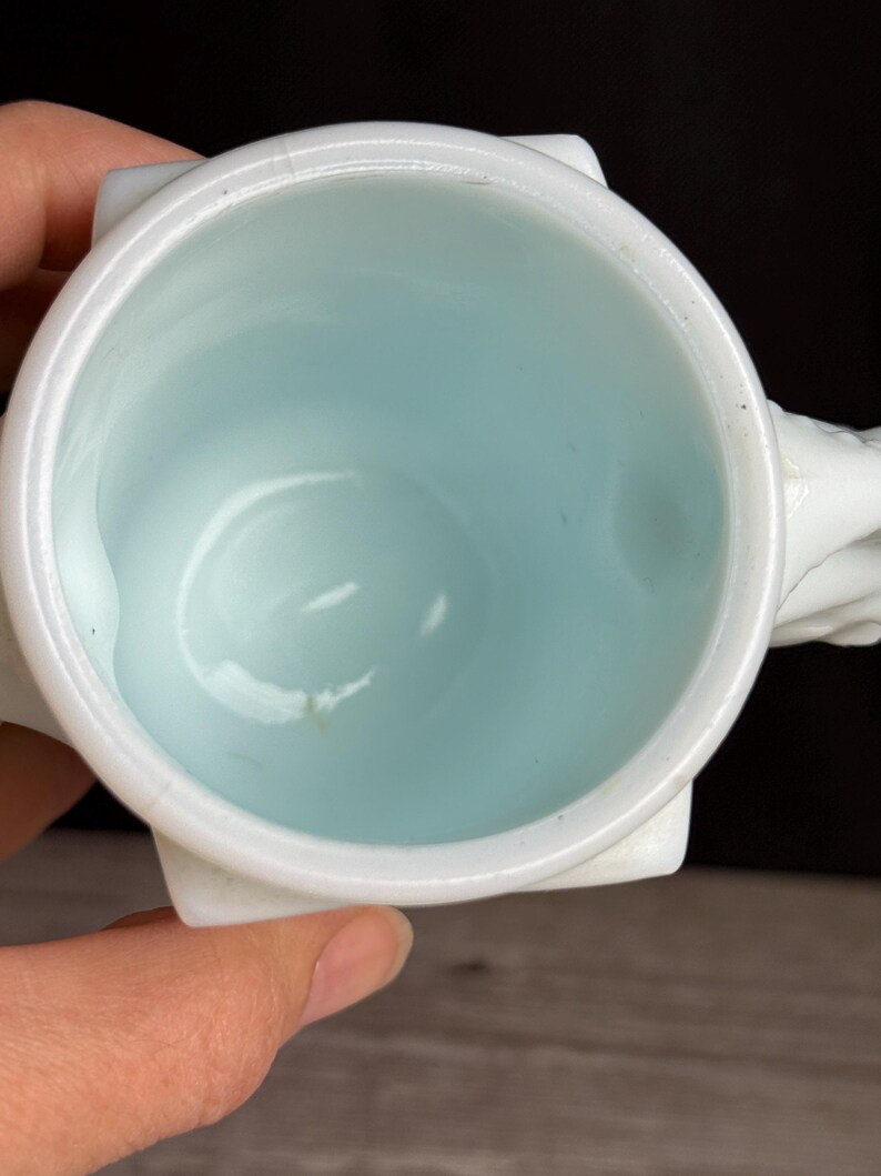 May include: A white ceramic mug with a light blue interior. The mug has a square base and a rounded rim. The handle is white and appears to be sculpted. The mug is set against a dark background.