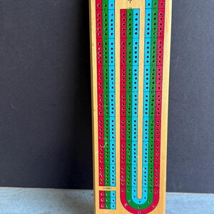 May include: Wooden cribbage board with red, green, and blue tracks. The board has the text "THE U.S. PLAYING CARD CO." and a start and finish area. The board is rectangular and has numerous holes for pegs.