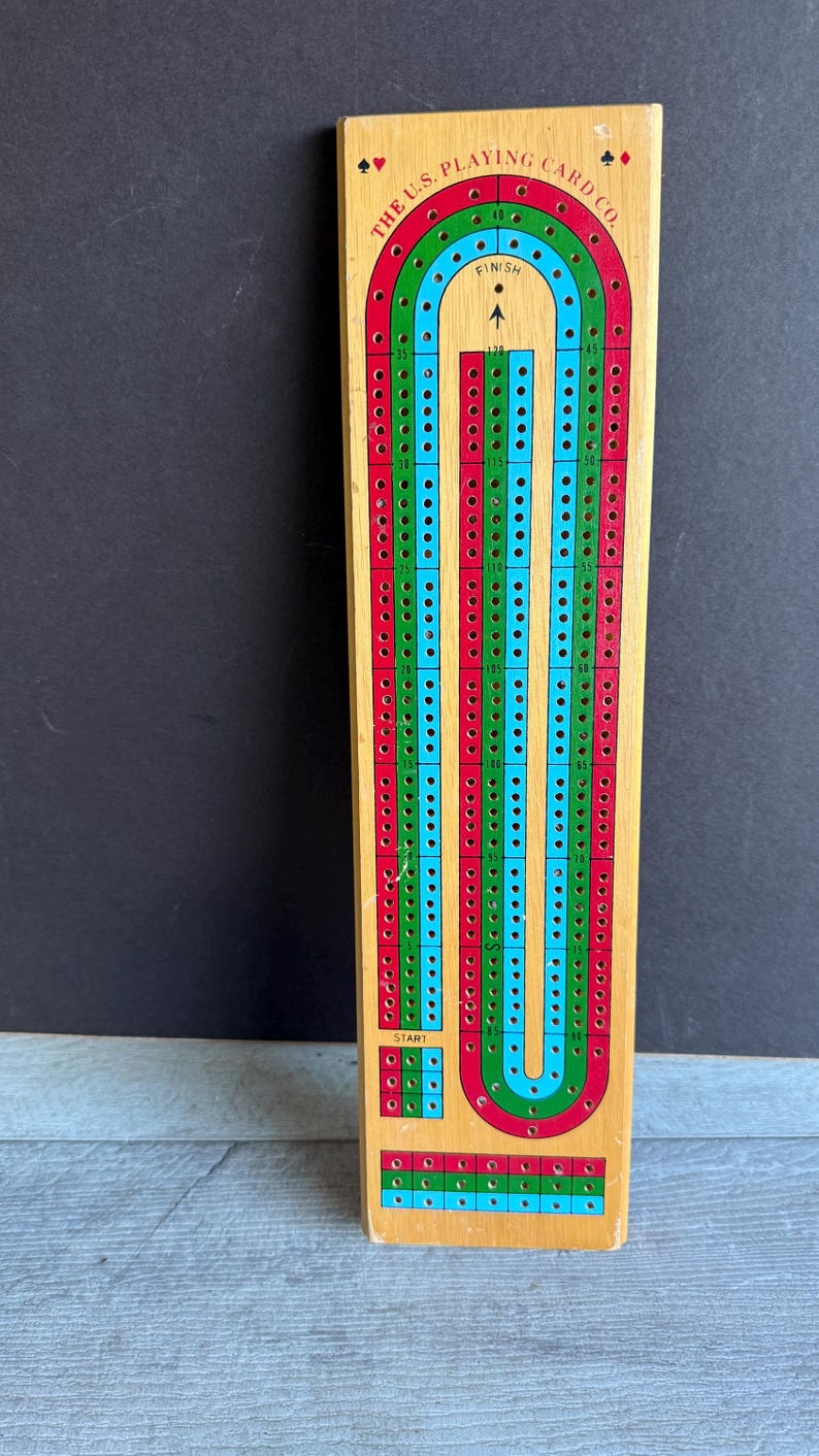 May include: Wooden cribbage board with red, green, and blue tracks. The board has the text "THE U.S. PLAYING CARD CO." and a start and finish area. The board is rectangular and has numerous holes for pegs.