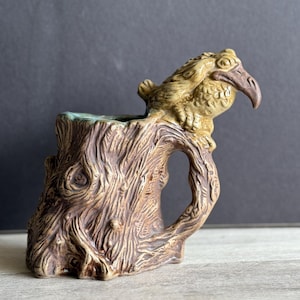 May include: A ceramic mug shaped like a tree stump with a bird perched on the top. The mug is brown and green with a textured surface.