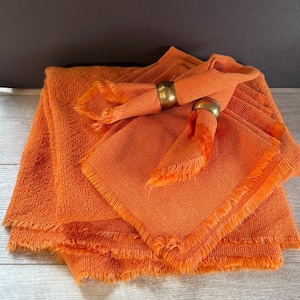 May include: Orange linen napkins with fringed edges and gold napkin rings. The napkins are folded and stacked, with two rings holding napkins in place. The color is a vibrant, warm orange.