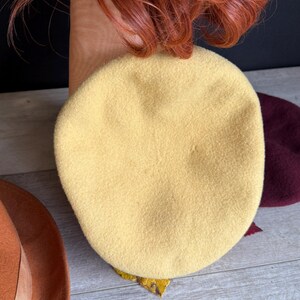 Puede incluir: Una boina de color amarillo claro hecha de tela suave. La boina es redonda y tiene una textura suave. Tambi&eacute;n se ven un sombrero marr&oacute;n con una cinta y una boina burdeos. La imagen tambi&eacute;n muestra cabello rojo.