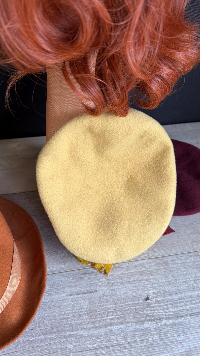 Puede incluir: Una boina de color amarillo claro hecha de tela suave. La boina es redonda y tiene una textura suave. Tambi&eacute;n se ven un sombrero marr&oacute;n con una cinta y una boina burdeos. La imagen tambi&eacute;n muestra cabello rojo.