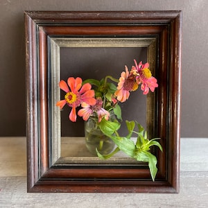 May include: A dark brown wooden picture frame with a decorative edge, holding a small glass jar with pink and yellow flowers and green leaves. The frame sits on a light gray surface.