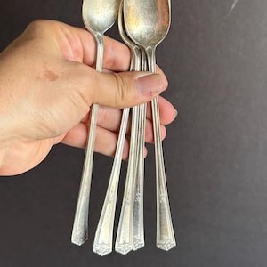 May include: A collection of vintage silver spoons. The spoons have an oval bowl and a long, slender handle with a decorative design at the end. The spoons are held in a hand against a dark background.