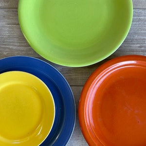 May include: Three ceramic plates, one green, one blue with a yellow plate inside, and one orange. The plates are on a wooden surface.